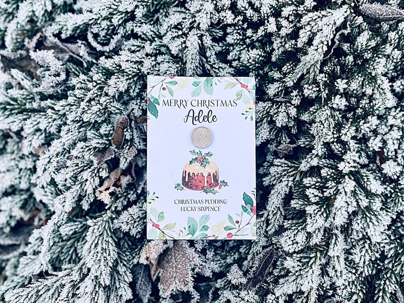 Festive Christmas greetings cards, each with a sixpence coin attached, displayed on a wooden surface with holly, berries, and a cozy fireplace and Christmas tree in the background.