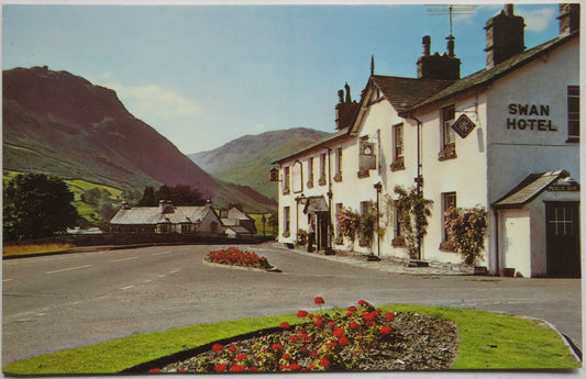 A vintage postcard-style photograph of the Swan Hotel in a mountain village, with white buildings, dark roofs, a paved road, flower beds, and green hills in the background under a blue sky.