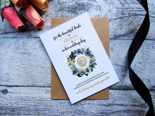 Personalised bride wedding card with blue floral wreath and real lucky sixpence, shown with kraft envelope and ribbon on a wooden background.